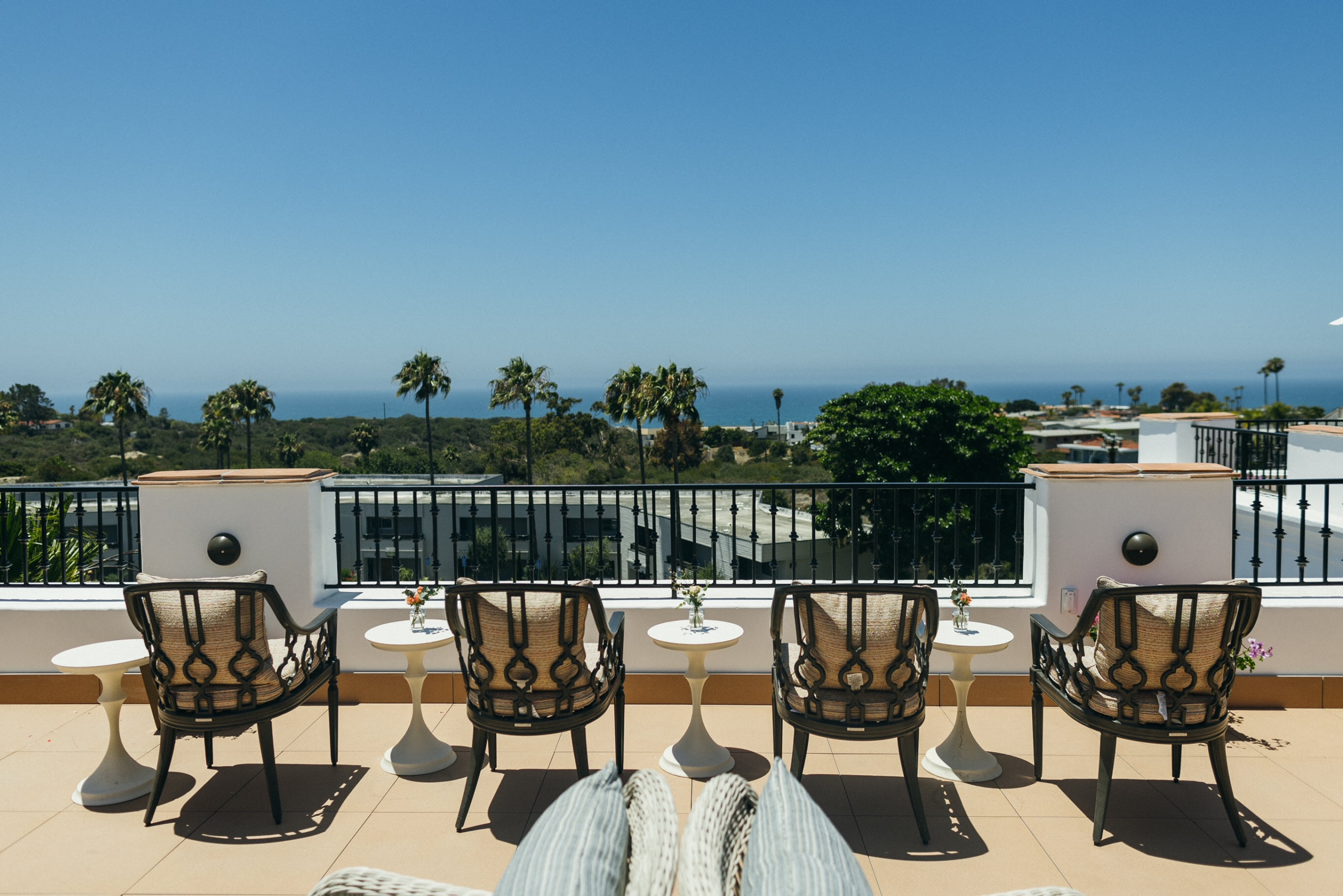 Outdoor patio chairs arranged along a terrace at Raya's Paradise with clear views of the Pacific Ocean, palm trees and the San Clemente coastline.