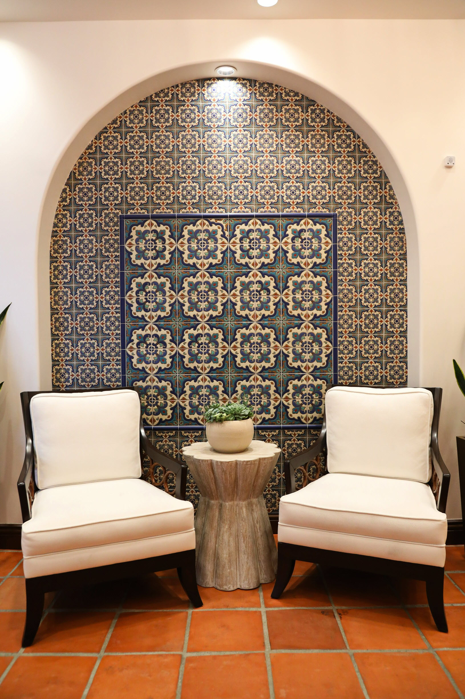 A pair of cushioned chairs sit beside a small table in front of a decorative Spanish tile alcove that adds warmth and character to the space.
