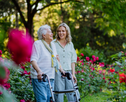 Younger woman accompanies an older adult using a walker through a flower garden, illustrating family caregiving and senior mobility support.